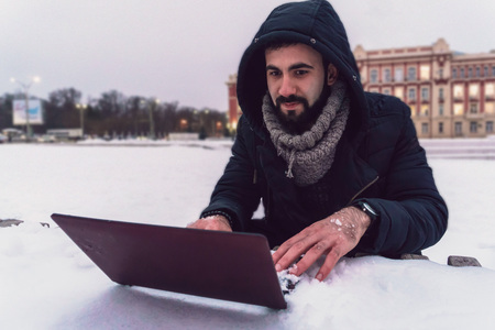 A young guy with a beard is working in the cold in the snow in the winter for a laptopの写真素材