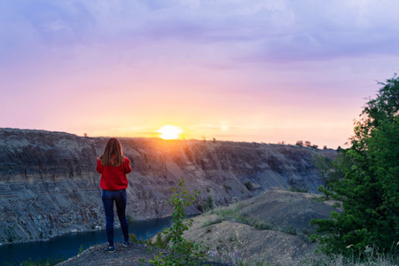 A young beautiful girl with a good figure in a red warm sweatshirt is standing on top of a hill above a blue lake and meets the gaze of dawnの写真素材