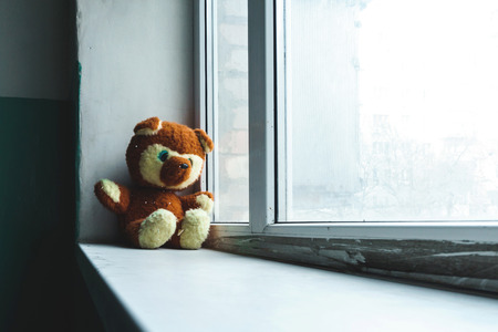Abandoned children's toy teddy bear unhappy child sits on the sills alone in the entrance and looks out the windowの写真素材