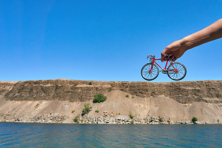 red metal toy bike in the hands against the background of a cliff in the distance with an optical illusion of a big bike on a clear sunny day in summer while travelingの写真素材