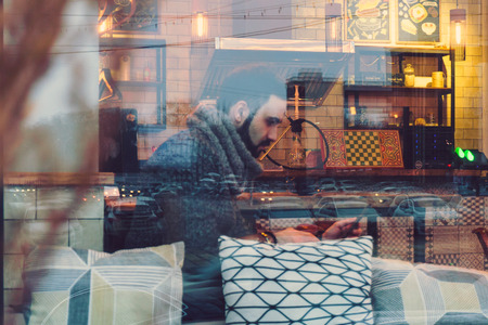 a young guy with a beard in a warm sweater works behind a laptop in a cozy cafe with pillows behind...