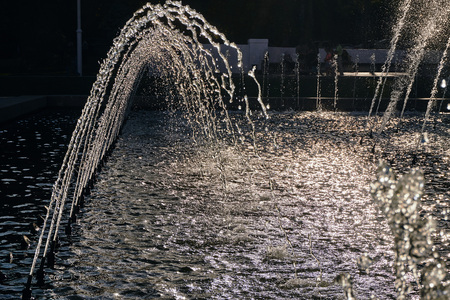 water jets of a large fountain shine in the rays in the central city park of the city in the air on a sunny summer dayの写真素材