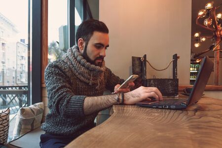 young guy with a beard in a sweater works at a computer in a cozy warm cafeの写真素材