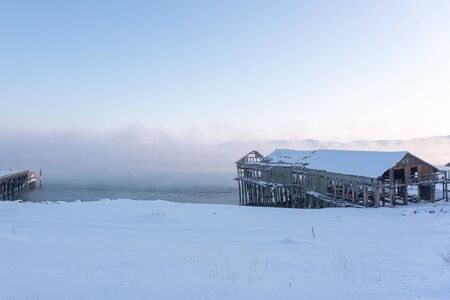 old wooden pier for large ships on the river bank covered in thick white fog from the water at dawn on a frosty winter day outside the Arctic Circleの写真素材