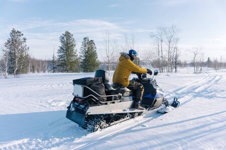 young guy in a yellow warm jacket sits on his snowmobile and rides through an empty snowy meadow surrounded by trees on a frosty winter dayの写真素材