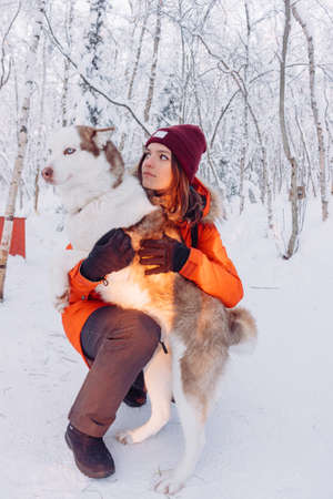 girl in a bright orange jacket sits and plays and hugs with a husky breed dog in the winter forest after snowfall during frost in the rays of the bright sunset sunの写真素材