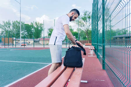 young guy in shorts on the sports ground parses a sports bag before starting a workout on a bench on a warm summer dayの写真素材