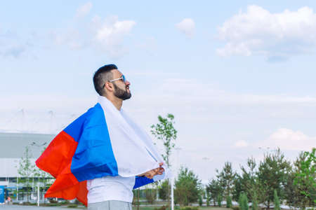 young guy with a stretched Russian flag on the site near a large stadium on a hot summer day before...