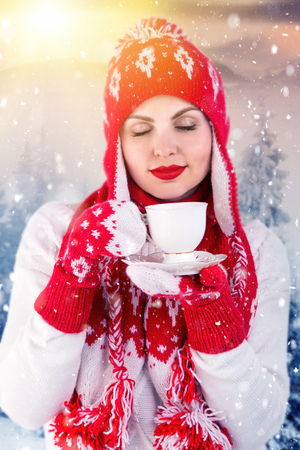 A young woman in a snowy forest drinking from a Cup of tea or coffee. Knitted hat, scarf and mittens.の写真素材
