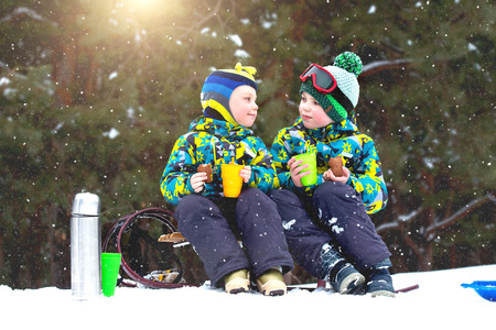 Two brothers drinking hot tea in a snowy forest after sledging. Winter fun for Christmas vacation.の写真素材