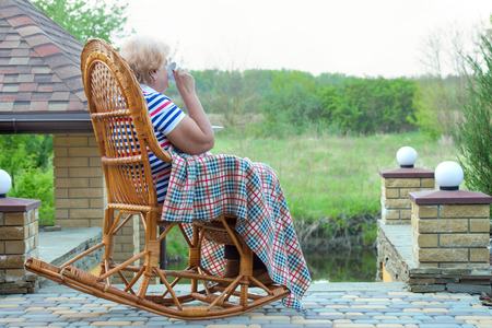 An elderly woman sits in a wicker rocking chair and a cup of hot tea.Relax in a country house.の写真素材