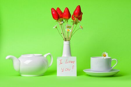 Berry bouquet. A bouquet of strawberries in a white vase and a small card on a green background. Cup of tea with chamomile and teapot.の写真素材