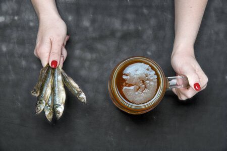 A woman holds a mug of cold beer and salted fish on a black wooden background.の写真素材