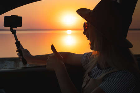 Young woman looking out car window of the sunset on the sea and and photographed on the phone.の写真素材
