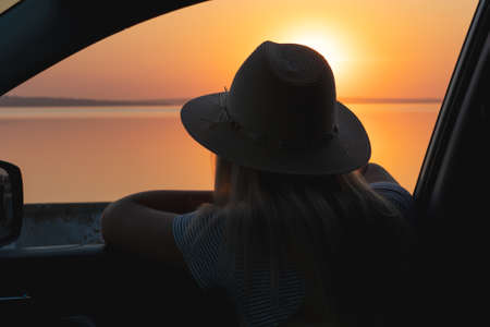 A young woman looks out the car window at the sunset on the sea.