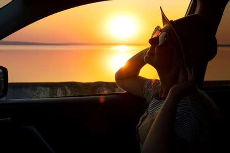 A young woman looks out the car window at the sunset on the sea.の写真素材