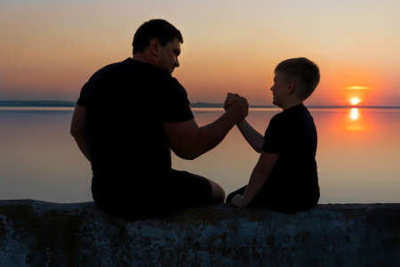 Strong friendship between father and son.Father and son sitting on the promenade and watch the sunset on the river.の写真素材