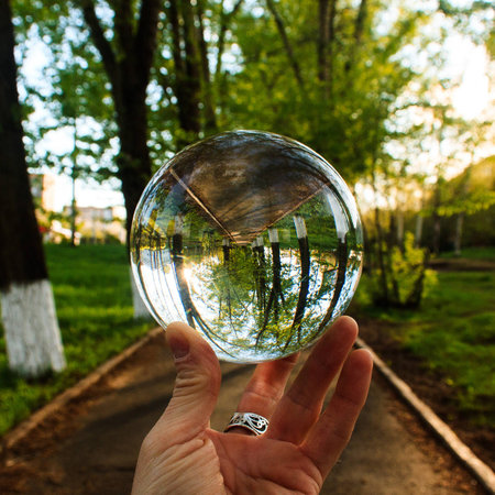 Arm holding the big transparent glass ball on the finger tips. Background is outdoor on the park...