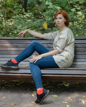 A red-haired girl sitting on a bench in an autumn park in a Golden jacket, blue jeans, red socks and black sneakersの写真素材