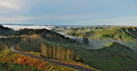 Road leading through the green hills with clouds rolling over them during the sunrise on North Island of New Zealand.の写真素材
