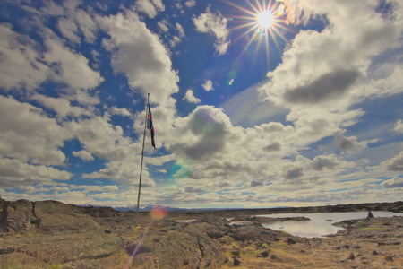 Icelandic flag on lava field with a lake nearby on backcountry hiking trail to Askja with clouds on sky and direct sunlight with some sun bokeh, interior highlands of Iceland.の写真素材