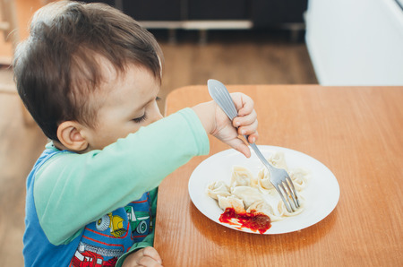 A hungry child is eating dumplings in the kitchen sitting in a chair in a white t-shirtの写真素材