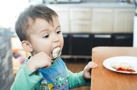 A hungry child is eating dumplings in the kitchen sitting in a chair in a white t-shirtの写真素材