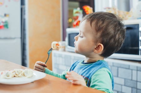 A hungry child is eating dumplings in the kitchen sitting in a chair in a white t-shirtの写真素材