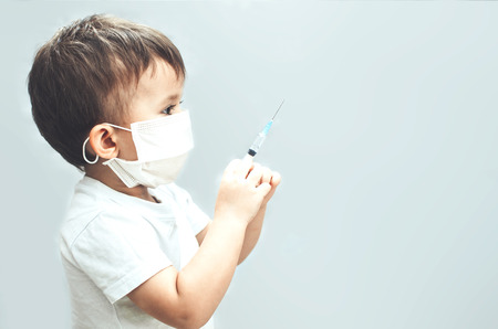 Child in a mask of the doctor with syringe in hand on gray light background, presents himself as a doctorの写真素材