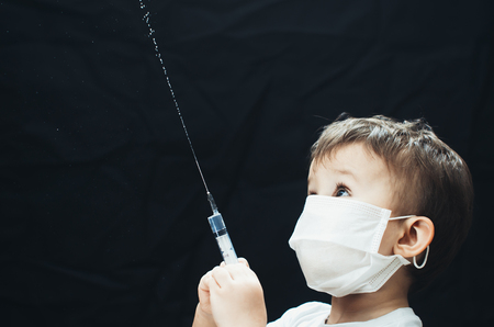 The child in the mask of the doctor with syringe in hands sprinkles, pours on a black backgroundの写真素材