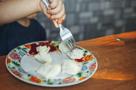 A hungry child is eating dumplings in the kitchen sitting in a chair in a white t-shirtの写真素材