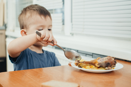 Child in the kitchen eating mashed potatoes, spoon, next to the plate is a piece of chicken meatの写真素材