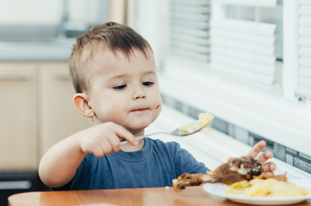 Child in the kitchen eating mashed potatoes, spoon, next to the plate is a piece of chicken meatの写真素材