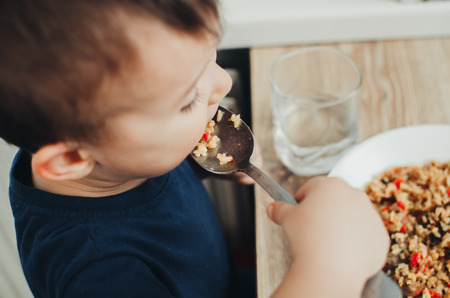 beautiful cute baby eats rice with a spoon in the kitchen, very funの写真素材