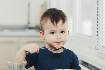 beautiful cute baby eats rice with a spoon in the kitchen, very funの写真素材