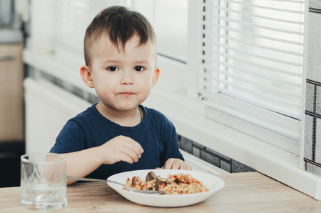 beautiful cute baby eats rice with a spoon in the kitchen, very funの写真素材