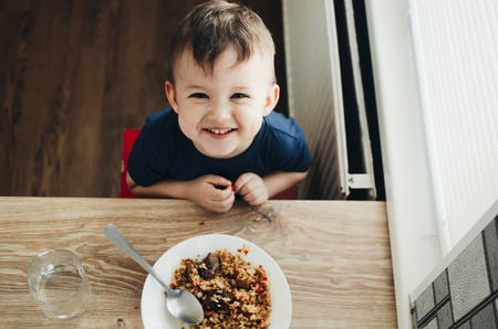 beautiful cute baby eats rice with a spoon in the kitchen, very funの写真素材