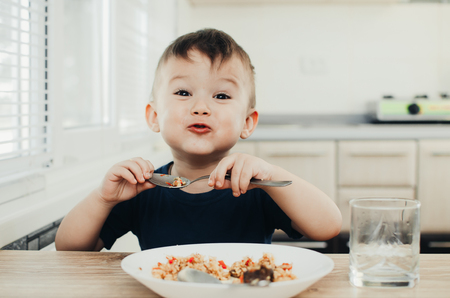 beautiful cute baby eats rice with a spoon in the kitchen, very funの写真素材