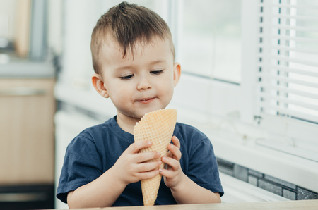 A child in a dark-blue t-shirt in the bright kitchen eating a waffle ice cream cone in the summer houseの写真素材
