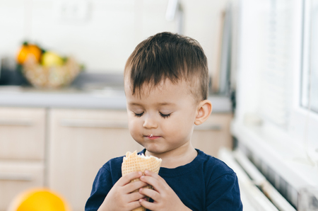 A child in a dark-blue t-shirt in the bright kitchen eating a waffle ice cream cone in the summer houseの写真素材