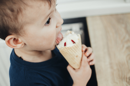 A child in a dark-blue t-shirt in the bright kitchen eating a waffle ice cream cone in the summer houseの写真素材