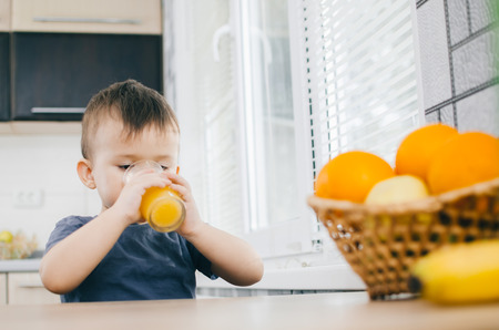 the child in the kitchen, drinking orange juice, close on background of fruitsの写真素材