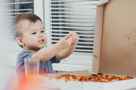 the little boy eats a huge harmful pizza himself in the kitchen and drinks juice, very fat and harmfulの写真素材