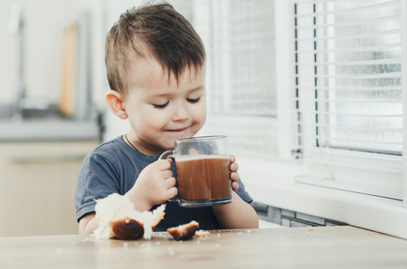 a child in the kitchen drinks hot chocolate and eats a sweet bunの写真素材