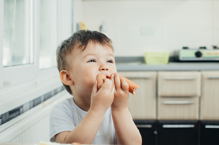 handsome boy shaggy and shkod eats sausage in the kitchen in a white t-shirt in the afternoonの写真素材