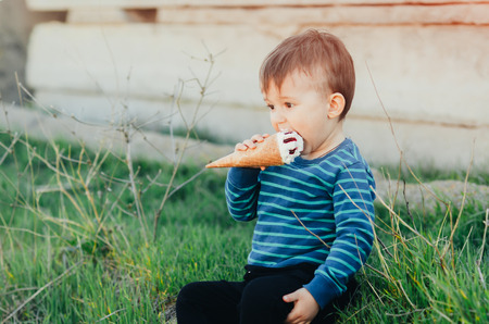 Cheerful child in a blue sweater eating ice cream on a background of grass, outdoors