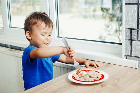 Hungry baby eating dumplings in the kitchen sitting on a chair in a blue t-shirtの写真素材