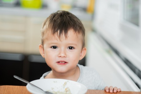 a charming child in a t-shirt in the kitchen eats oatmeal very greedilyの写真素材