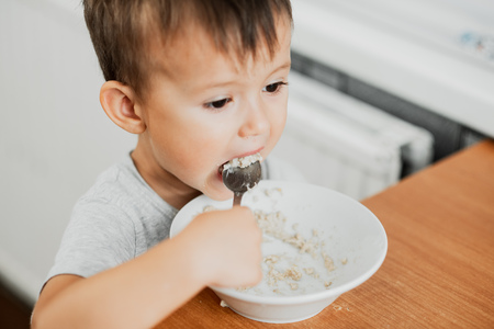 a charming child in a t-shirt in the kitchen eats oatmeal very greedilyの写真素材