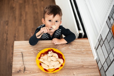 Hungry child eating dumplings in the kitchen, sitting at the table in a gray jacketの写真素材